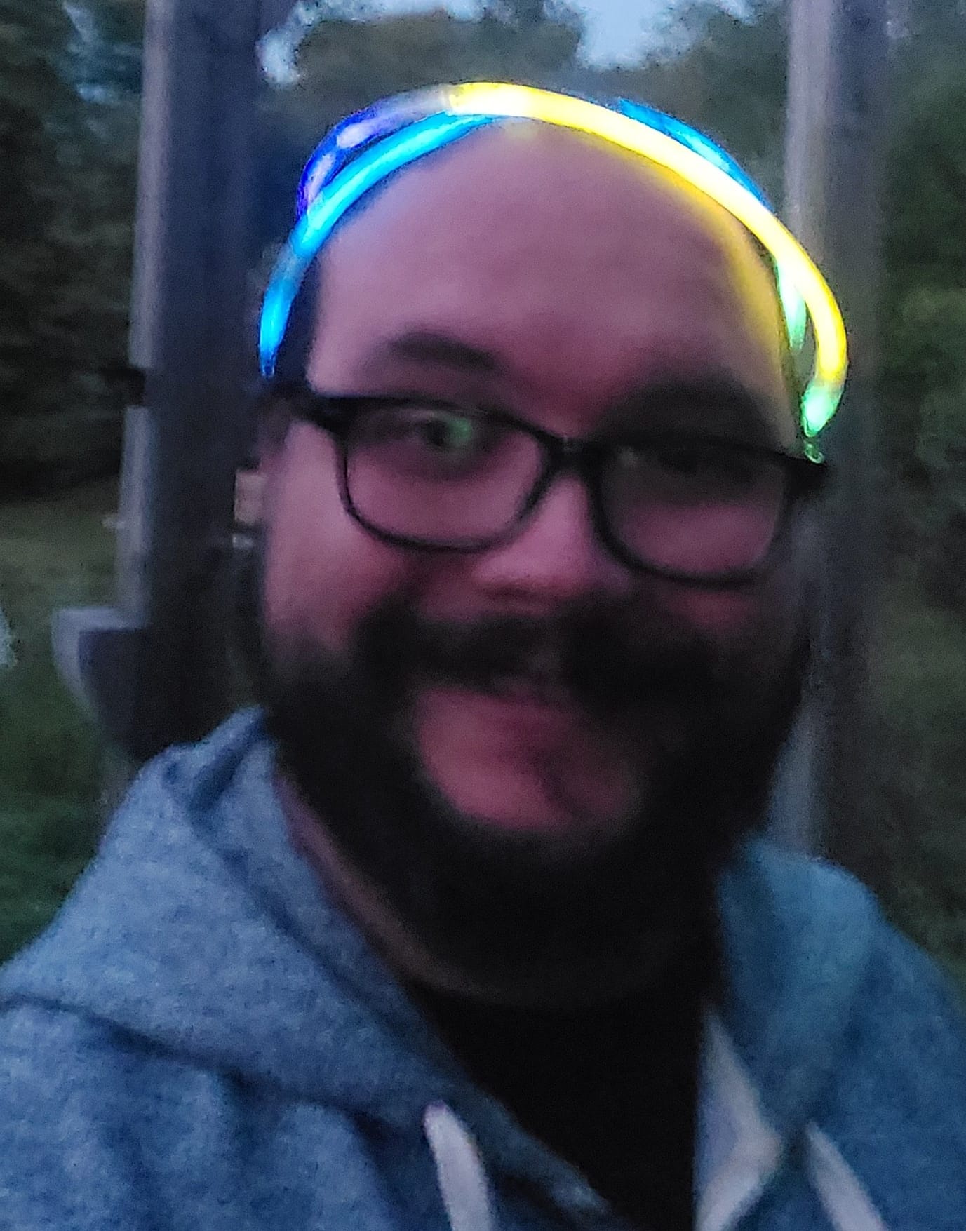 A man with facial hair wears a grey hoodie and a glow-stick circlet